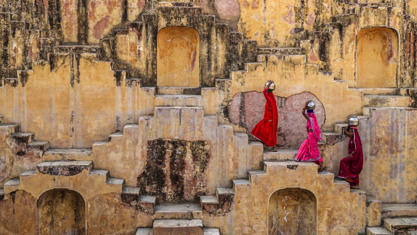 Indian women carrying water from stepwell near Jaipur, Rajasthan, India. Women and children often walk long distances to bring back jugs of water that they carry on their head. 
Stepwells are wells in which the water may be reached by descending a set of steps.